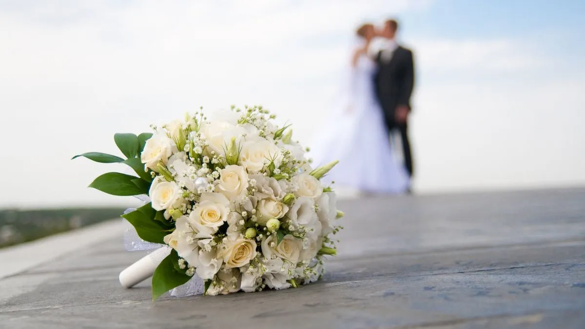 Primo piano di un bouquet da sposa con rose bianche e fiori di gypsophila su un patio, con la silhouette sfocata di sposi che si baciano sullo sfondo.